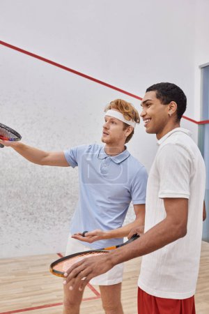 happy interracial players in sportswear smiling and looking away inside of squash court, racquets