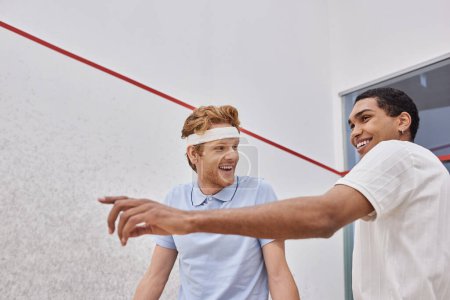 happy interracial players pointing with racquets and looking away inside of squash court, friends