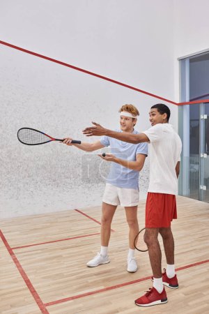 happy interracial male friends pointing with racquets and looking away inside of squash court