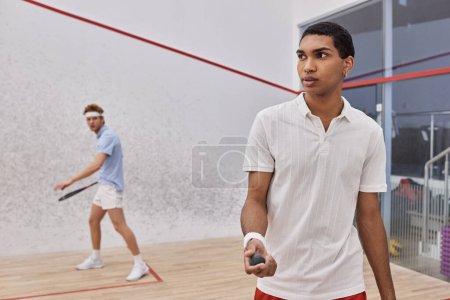 young african american man holding squash ball and playing with redhead friend inside of court
