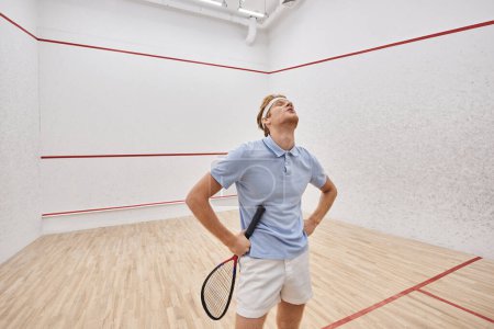 exhausted redhead man in headband and sportswear breathing heavily after playing squash in court
