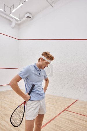 hombre agotado en diadema y ropa deportiva respirando pesadamente después de jugar squash dentro de la corte