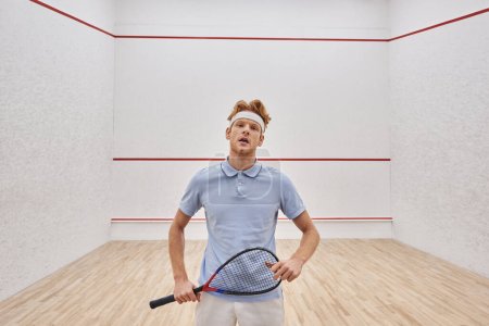hombre cansado en diadema y ropa deportiva respirando pesadamente después de jugar squash dentro de la corte