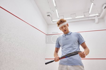 hombre cansado en diadema y ropa deportiva respirando pesadamente y mirando raqueta de squash dentro de la corte