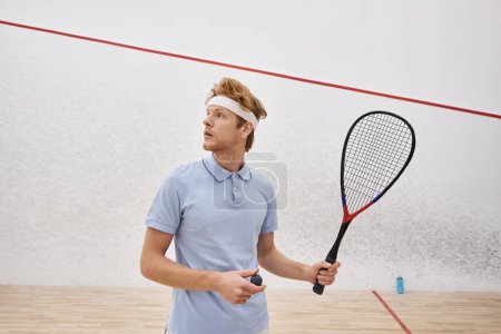 redhead man in sportswear holding squash ball and racquet while standing inside of indoor court