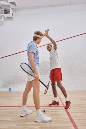 cheerful multicultural friends in sportswear giving high five after playing squash in court