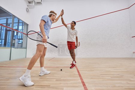 two happy multicultural friends in sportswear giving high five after playing squash in court