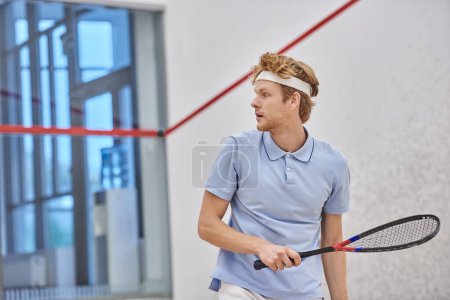 athletic redhead sportsman holding racquet while playing squash inside of court, training shot