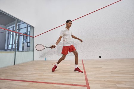 african american sportsman in red sporty shorts holding racquet while playing squash inside of court