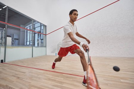 sportif afro-américain en short de sport rouge tenant la raquette tout en jouant au squash, motion shot
