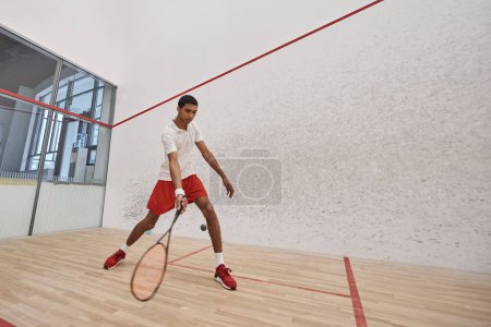 junger afrikanisch-amerikanischer Mann in roten, sportlichen Shorts mit Schläger beim Squash-Spiel, Bewegungsaufnahme