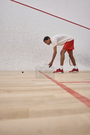 african american man in red sporty shorts holding racquet and picking up squash ball inside of court