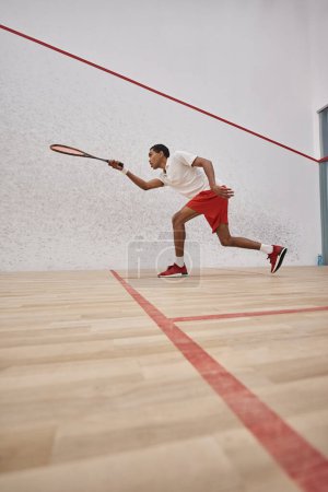 energetic african american sportsman in shorts holding racquet and playing squash inside of court