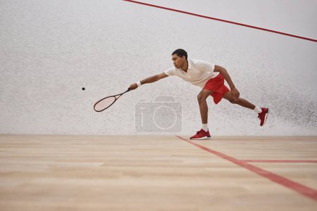 african american sportsman in red shorts holding racquet and playing squash inside of court, sport