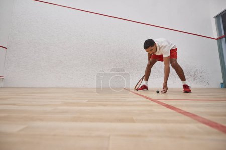 african american man in red shorts holding racquet and picking up squash ball inside of court