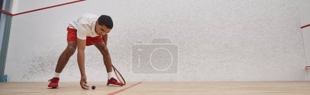 afro-américain en short rouge tenant raquette et ramassant balle de squash au tribunal, bannière