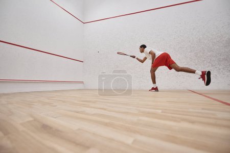 african american sporty man in red shorts holding racquet while playing squash inside of court