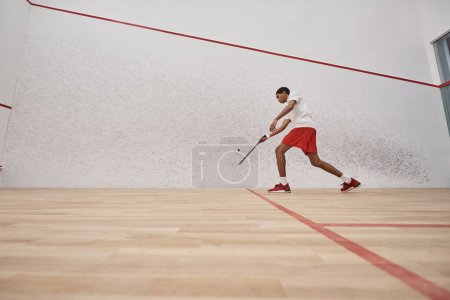 african american young man in red shorts holding racquet while playing squash inside of court
