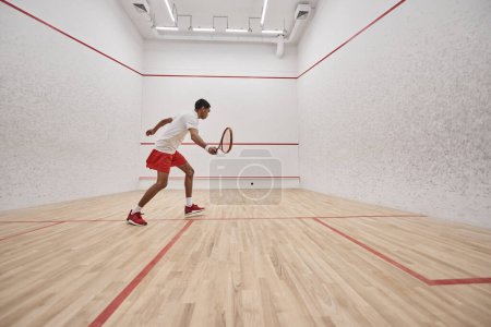 african american sportsman in sports shorts holding racquet while playing squash inside of court