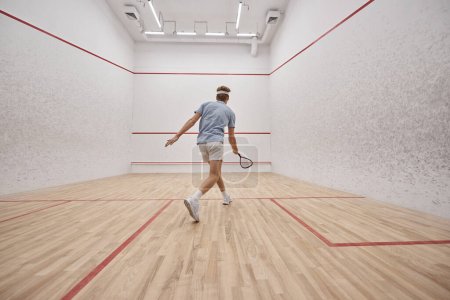 athletic and redhead sportsman holding racquet while playing squash inside of court, motion shot