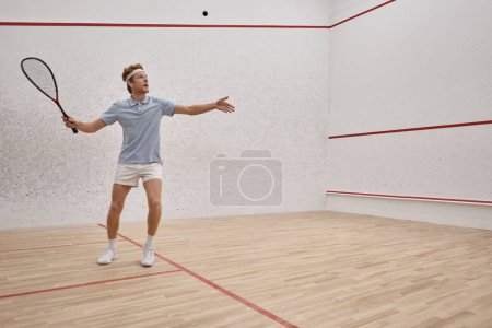 athletic redhead man in sportswear holding racquet and playing squash inside of court, motion shot