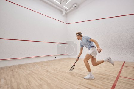athletic and redhead sportsman with racquet playing squash inside of court, motion shot