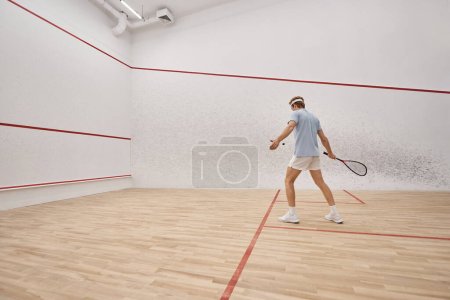 sporty and redhead man holding racquet while playing squash inside of court, motion shot