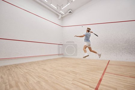 energetic and redhead sportsman holding racquet and playing squash inside of court, motion shot