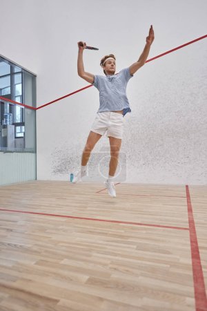 foto en movimiento, deportista divertido celebración de raqueta y saltar mientras juega squash dentro de la cancha