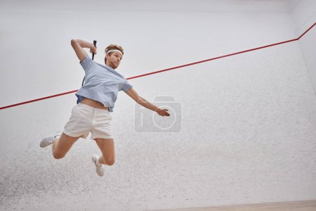 tiro en movimiento, deportista motivado sosteniendo raqueta y saltando mientras juega squash dentro de la cancha