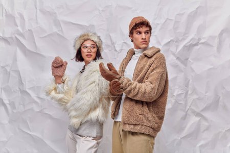 Téléchargez les photos : Couple interracial en fausse fourrure chaude vestes regardant loin sur fond blanc texturé, style d'hiver - en image libre de droit