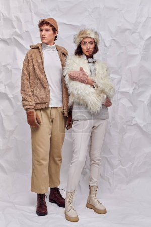 full length of multiethnic couple n fashionable winter attire on white crumpled paper backdrop