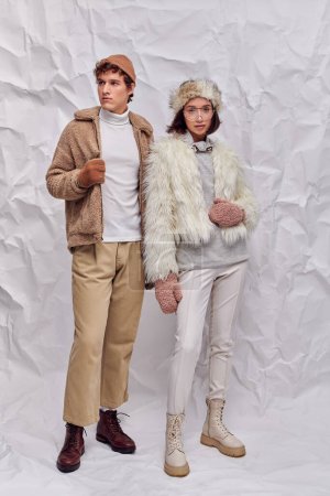 asian woman in faux fur jacket and mittens looking at camera near trendy man on white paper backdrop