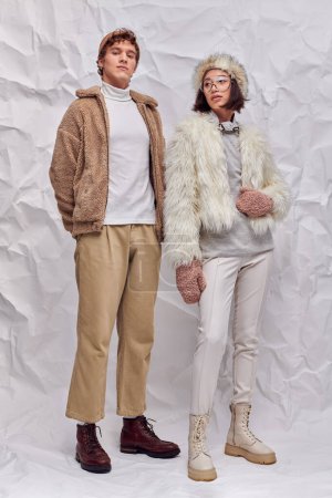 hombre de moda mirando a la cámara cerca de mujer asiática en chaqueta caliente y manoplas sobre fondo texturizado blanco