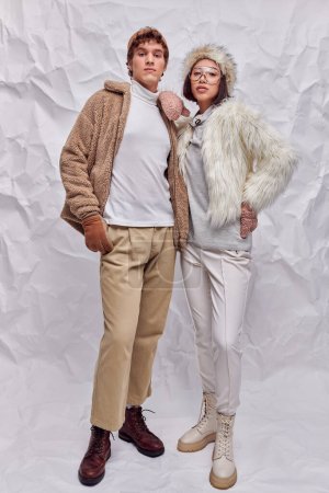 young asian model in warm attire posing with hand on hip near trendy man on white textured backdrop