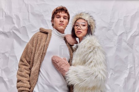 stylish man and asian woman in faux fur jacket and mittens looking at camera on white paper backdrop
