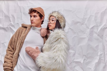 fashion lookbook concept, interracial couple in winter wear looking away on white crumpled backdrop