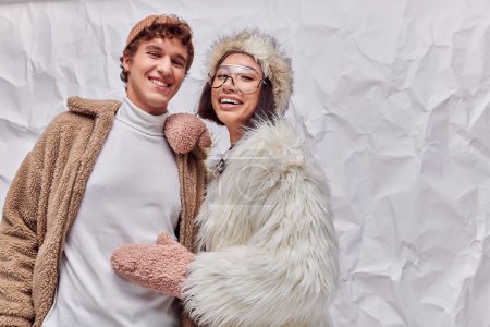 joyful interracial couple in fashionable warm outwear on white textured backdrop, winter fashion