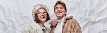 happy interracial couple in stylish winter wear looking at camera on white textured backdrop, banner