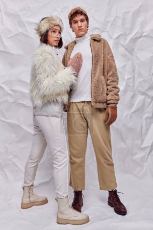 young trendy man looking at camera near asian woman in warm cozy clothes on white crumpled backdrop