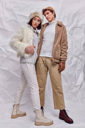 multiethnic couple in trendy winter clothes looking away on white textured backdrop, seasonal style