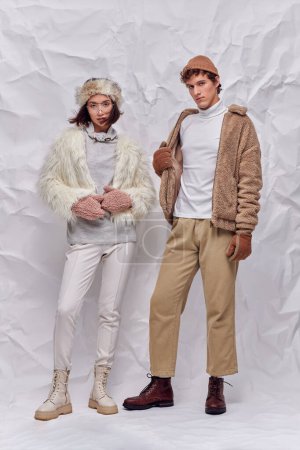 interracial couple in hats, warm gloves and faux fur jackets standing on white crumpled backdrop
