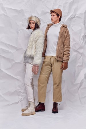 in voller Länge trendige interrassische Models in winterlichen Outfits vor weiß strukturiertem Hintergrund, moderner Trend