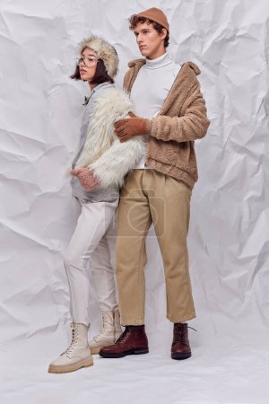 full length of multiethnic couple in worm cozy outfits looking away  on white textured backdrop