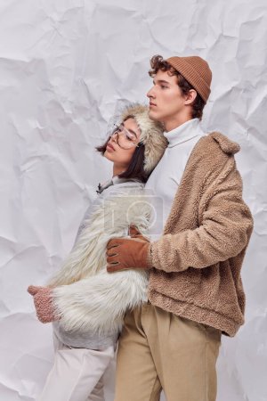 trendy man in warm clothes hugging young asian model and looking away on white crumpled backdrop