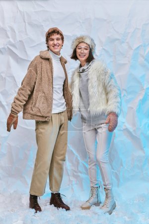 aufgeregtes gemischtes Paar in warmer stylischer Kleidung, die im Atelier auf Schnee steht, Wintermode