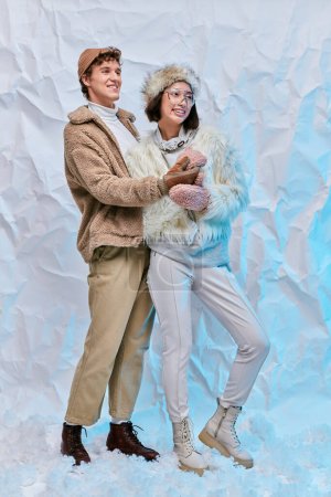 pareja interracial sin preocupaciones en elegante atuendo de invierno mirando hacia otro lado en la nieve sobre fondo de textura blanca