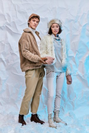 trendy man holding hand of asian woman in mittens standing on snow on white textured background