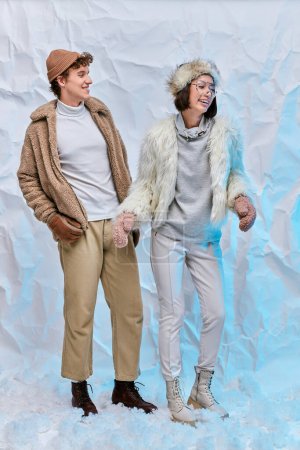 fashionable multiethnic couple in cold-weather attire laughing on snow in studio, winter happiness