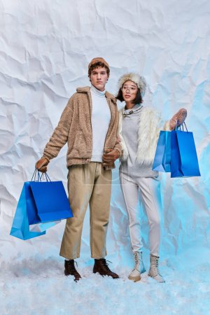 fashionable interracial couple with blue shopping bags on white snow in studio, winter purchases